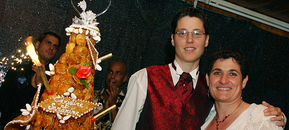 Couple de mariés souriant devant une pièce montée décorée, ambiance festive lors d'un mariage, gâteau personnalisé avec inscription 'Christelle Sandrine', moment de joie entouré de proches et d'animations lumineuses.